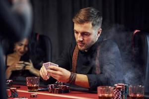 Elegant young people sits by table, playing poker in casino and checking their cards photo