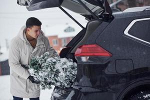 hombre en bata blanca poniendo abeto de navidad dentro de su automóvil foto