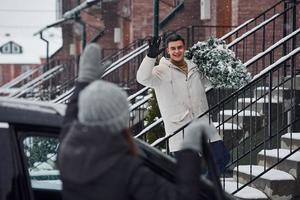 Man with christmas tree standing on the ladder of building and greeting female friend that is near car photo