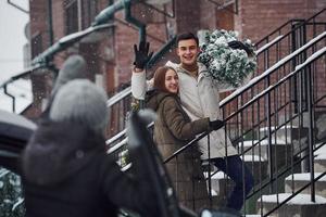 Young couple with christmas fir tree standing on the ladder of building and talking to female friend that near the car photo