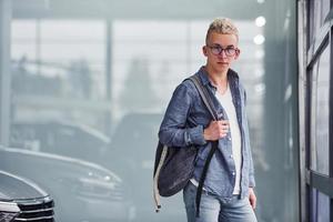 Young hipster guy in nice clothes stands indoors against grey background near modern car photo
