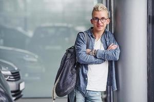 Young hipster guy in nice clothes stands indoors against grey background near modern car photo