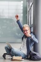 Happy hipster guy sitting indoors near grey background with laptop and books photo