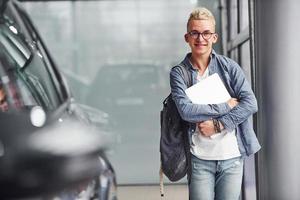 Young hipster guy in nice clothes stands indoors against grey background and near car photo