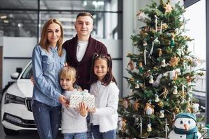 Happy family stands together in salon with car behind and christmas tree photo