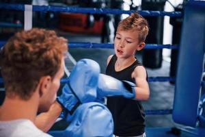 Young boxing coach is helping little boy in protective wear on the ring between the rounds photo
