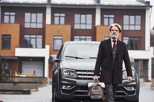 Fashionable senior man with gray hair and beard walking outdoors on the street near his car photo