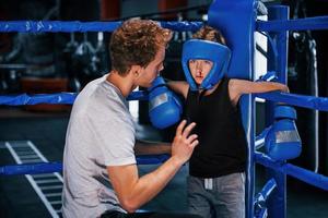 el joven entrenador de boxeo está ayudando a un niño pequeño con ropa protectora y con sangre debajo de la nariz en el ring entre las rondas foto