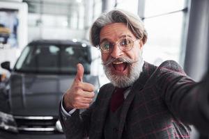 Stylish senior man in formal clothes and with beard takes selfie against modern new car. Showing thumb up photo