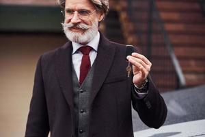 hombre mayor de moda con pelo gris y barba parado afuera en la calle cerca de su auto con llaves en la mano foto