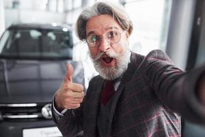 Stylish senior man in formal clothes and with beard takes selfie against modern new car. Showing thumb up photo