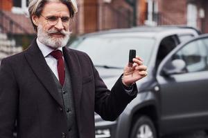 hombre mayor de moda con pelo gris y barba parado afuera en la calle cerca de su auto con llaves en la mano foto