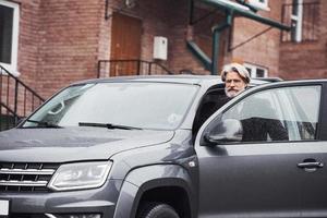 un anciano de moda con pelo gris y barba está al aire libre en la calle sentado en su coche foto