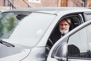 un anciano de moda con pelo gris y barba está al aire libre en la calle sentado en su coche foto