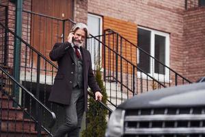 Fashionable senior man with gray hair and beard is outdoors on the street near his car photo