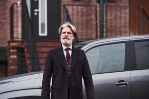 un anciano de moda con pelo gris y barba está al aire libre en la calle cerca de su coche foto