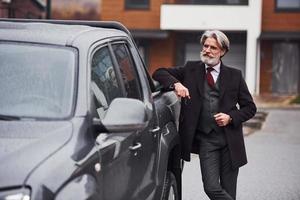 hombre mayor de moda con pelo gris y barba parado afuera en la calle cerca de su auto con llaves en la mano