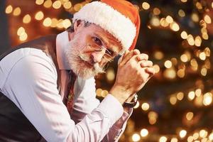 retrato de elegante anciano con cabello gris y barba en chaqueta y camisa blanca está en la sala de navidad decorada foto