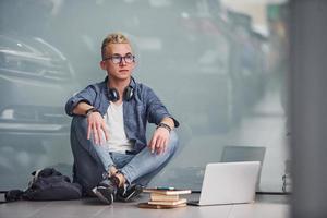 Young hipster guy sitting indoors near grey background with laptop and books photo
