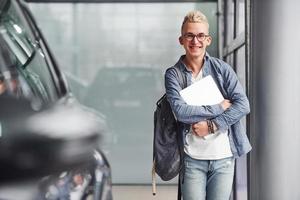 un joven hipster con ropa bonita se para en el interior contra un fondo gris y cerca del auto foto