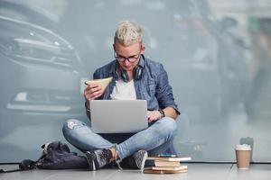 Young hipster guy sitting indoors near grey background with laptop and books photo