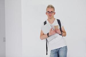 Student in casual clothes and with backpack stands indoors against white wall with cup of drink and laptop photo