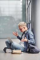 joven hipster sentado en el interior cerca de un fondo gris con laptop y libros foto