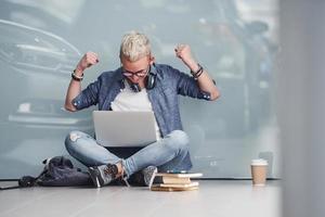 Young hipster guy sitting indoors near grey background with laptop and books photo