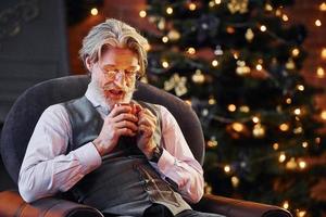 Portrait of stylish senior with grey hair and beard is in decorated christmas room with cup of drink photo