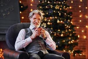 Portrait of stylish senior with grey hair and beard is in decorated christmas room with cup of drink photo