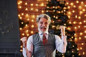 Portrait of stylish senior with grey hair and beard in jacket and white shirt is in decorated christmas room photo