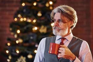 Portrait of stylish senior with grey hair and beard is in decorated christmas room with cup of drink photo