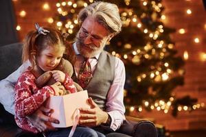 hombre mayor de moda alegre con pelo gris y barba con niña con caja de regalo foto