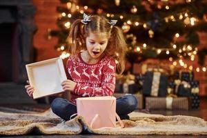 Cute little girl sits in the decorated christmas room opening gift box photo