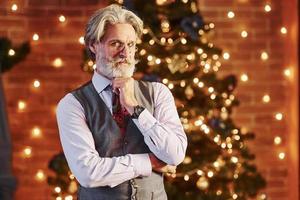 retrato de elegante anciano con cabello gris y barba en chaqueta y camisa blanca está en la sala de navidad decorada foto