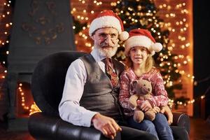 hombre mayor de moda alegre con cabello gris y barba sentado con una niña con sombreros de navidad y oso de peluche foto