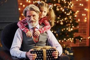 hombre mayor de moda alegre con pelo gris y barba con una niña pequeña con caja de regalo abrazándose unos a otros foto