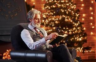 Portrait of stylish senior with grey hair and beard reading book in decorated christmas room photo