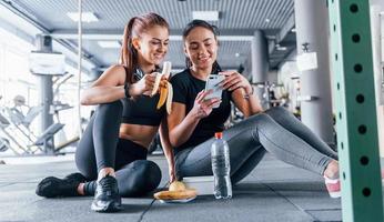 Two female friends in sportive clothes is in the gym earing fruits and using phone photo