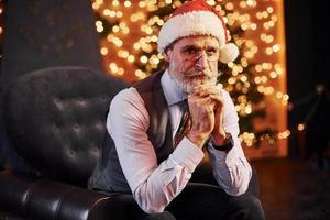 Portrait of stylish senior with grey hair and beard sitting in decorated room and in christmas hat photo