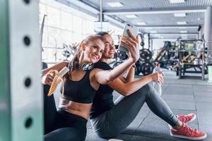 Two female friends in sportive clothes is in the gym earing fruits and taking a selfie photo