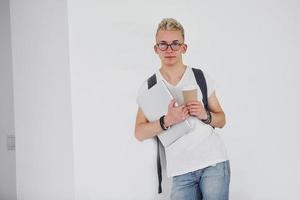 Student in casual clothes and with backpack stands indoors against white wall with cup of drink and laptop photo