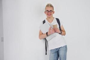 Student in casual clothes and with backpack stands indoors against white wall with cup of drink and laptop photo