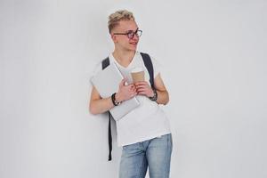 Student in casual clothes and with backpack stands indoors against white wall with cup of drink and laptop photo