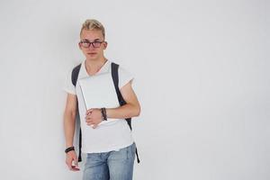 Student in casual clothes and with backpack stands indoors against white wall with laptop photo