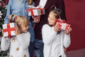 dos niñas se paran junto con mamá y papá en la habitación con un árbol de navidad con cajas de regalo foto