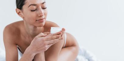 Young woman in underwear lying down in the studio against white background with beauty cream in hands photo