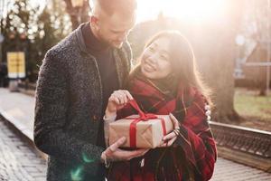 Beautiful sunlight. Happy multiracial couple together outdoors in the city with gift box in hands photo