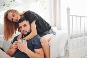 Young married couple with cup of drink have leisure together in bedroom at daytime photo