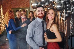 joven barbudo con su novia parados juntos contra sus amigos en una habitación decorada de navidad y celebrando el año nuevo foto